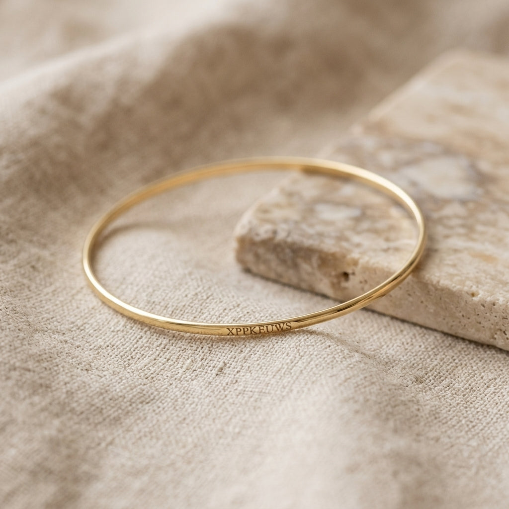 A thin gold bangle bracelet with the Lat & Lo Memory Key code engraved on the outside, resting on a textured beige linen cloth next to a stone