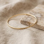 A thin gold bangle bracelet with the Lat & Lo Memory Key code engraved on the outside, resting on a textured beige linen cloth next to a stone