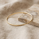 A thin gold bangle bracelet with the Lat & Lo Memory Key code engraved on the outside, resting on a textured beige linen cloth next to a stone