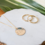 two gold rings inscribed with latitude longitude coordinates by Lat & Lo. Pictured next to a 16mm round coordinates disc necklace in gold-filled. by Lat & Lo. www.latandlo.com